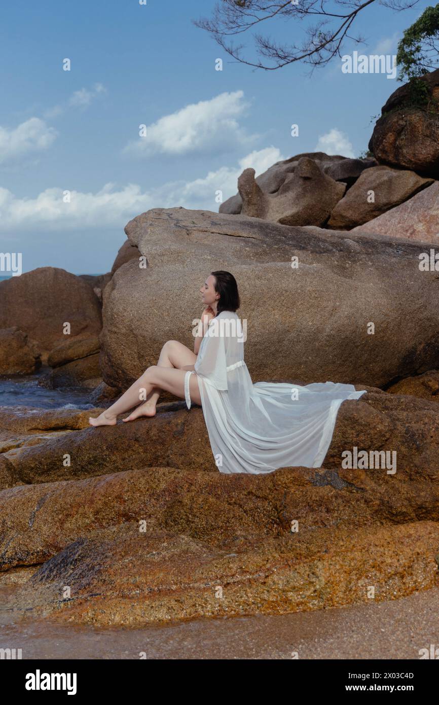 Woman in a flowing white dress enjoys a peaceful moment on coastal rocks, the sea softly lapping near her Stock Photo