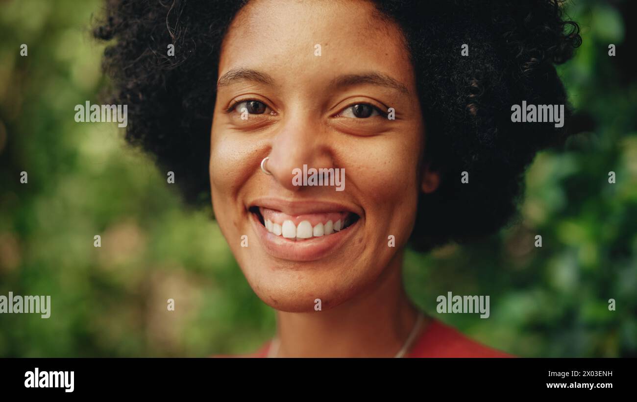 Close Up Portrait of a Happy Young Latina with Brown Eyes, Afro Hair and Nose Piercing Posing for Camera. Beautiful Diverse Multiethnic Black Hispanic Female Smiling on Green Nature Background. Stock Photo