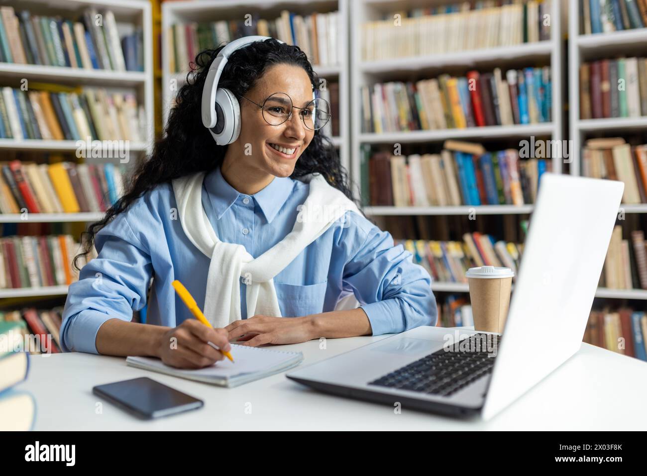 Focused student engages with online learning in a library setting, using headphones and taking notes alongside a cup of coffee. Stock Photo