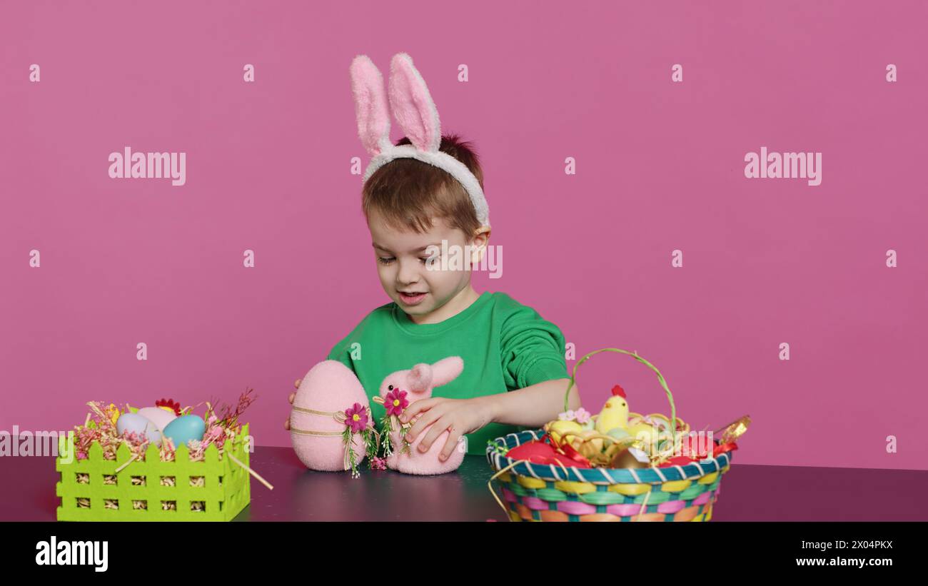 Joyful young kid playing around with festive painted decorations, showing a rabbit toy and a pink egg in front of camera. Smiling small boy with bunny ears having fun with ornaments. Camera A. Stock Photo