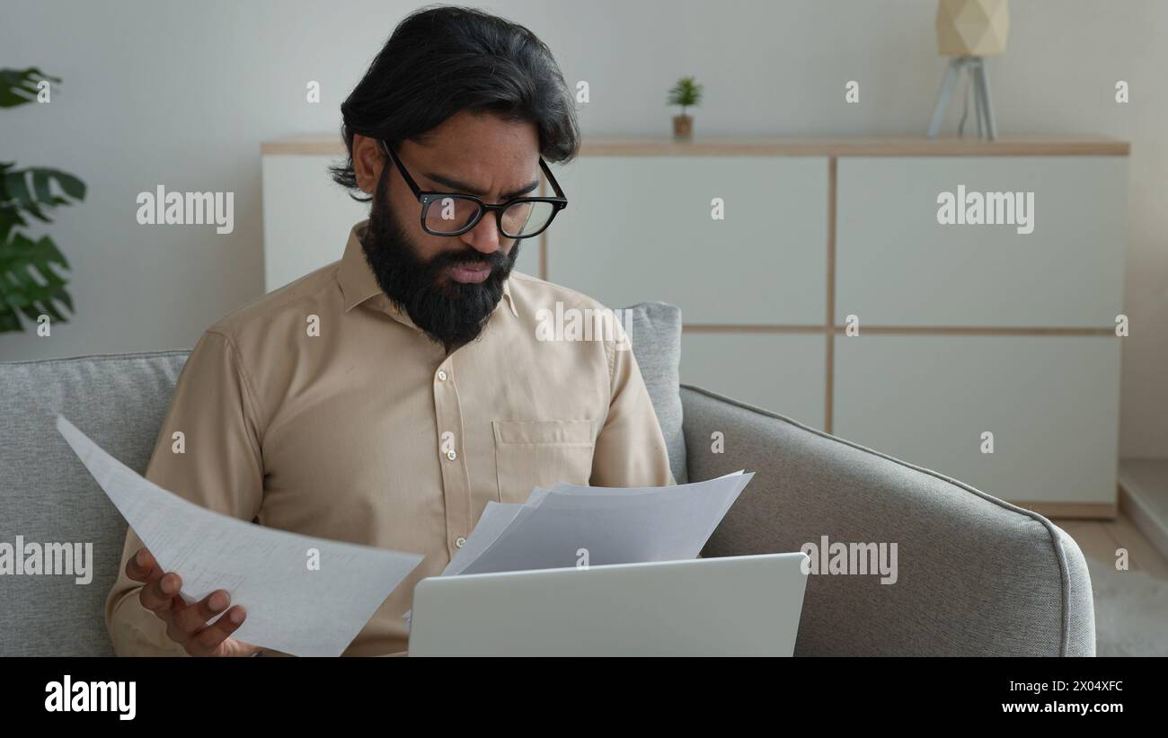 Anxious Arabian businessman freelancer on sofa reading documents pay bills at home managing finances taxes stressed man sorting out papers fees high Stock Photo