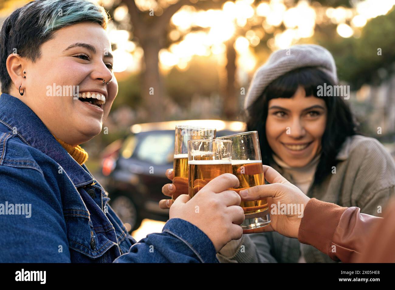 Non-binary or Gender-fluid person with a curvy figure shares a cheerful toast with a friend, smiling, during a relaxed outdoor gathering at golden hou Stock Photo
