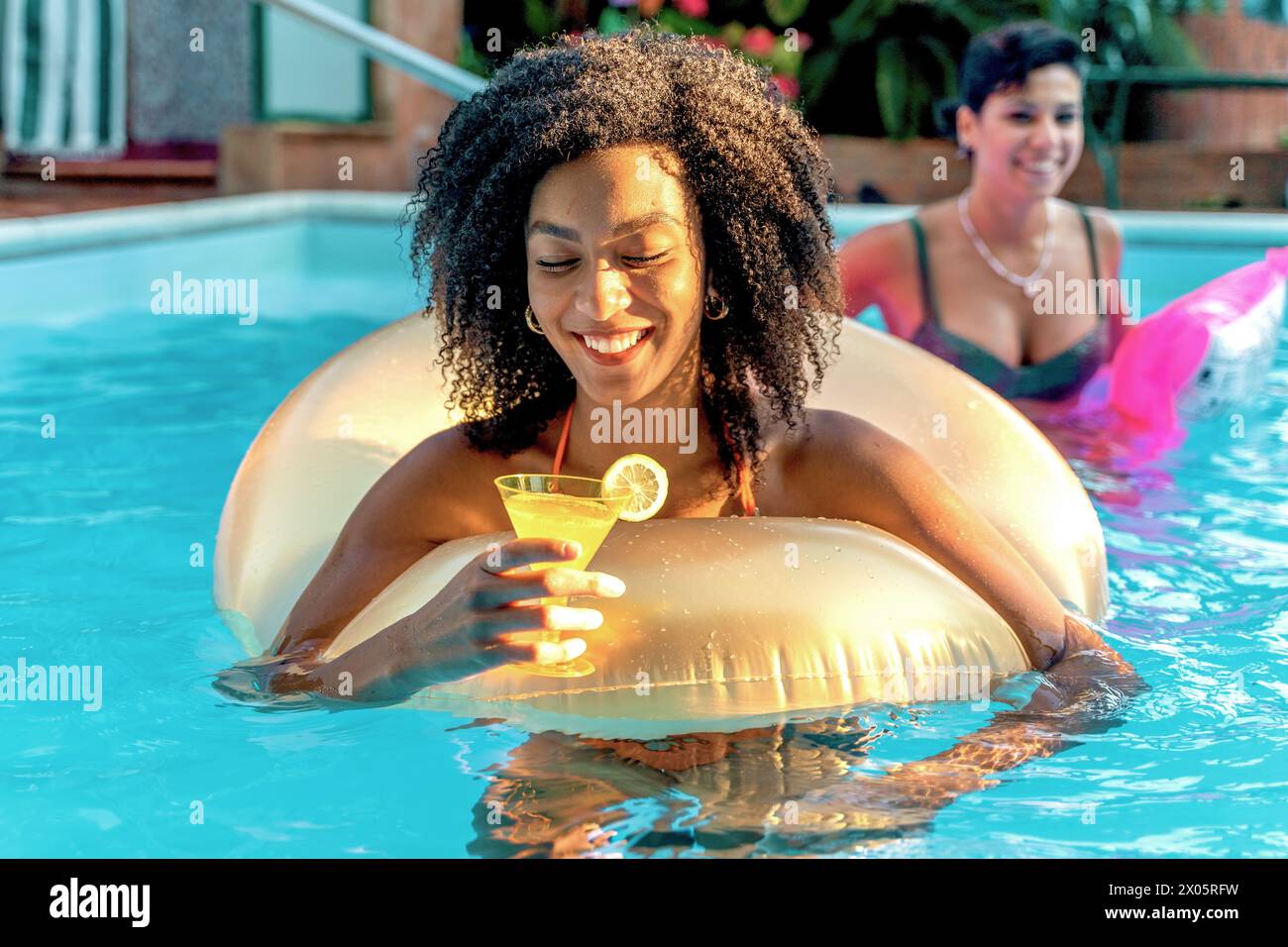 African descent woman with a bright smile relaxes on a golden inflatable ring in a pool, sipping a refreshing lemon cocktail on a sunlit day, with a f Stock Photo