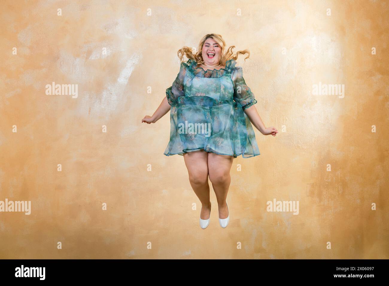Excited plus size woman in green dress leaping in joy against yellow background in studio Stock Photo