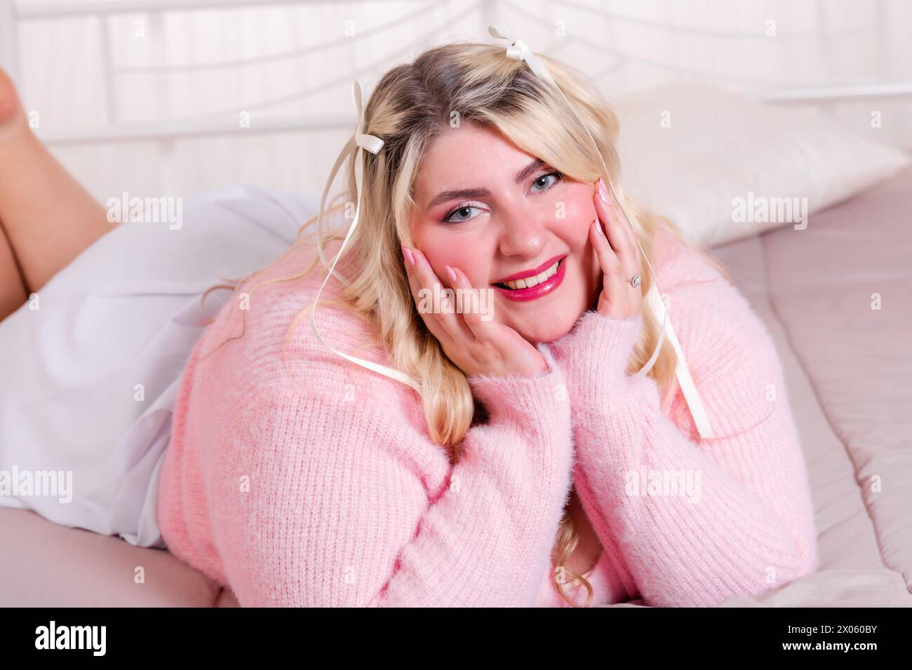 Smiling plus size young lady wearing casual outfit relaxing on comfortable bed with hands on chin and looking at camera in bright room Stock Photo