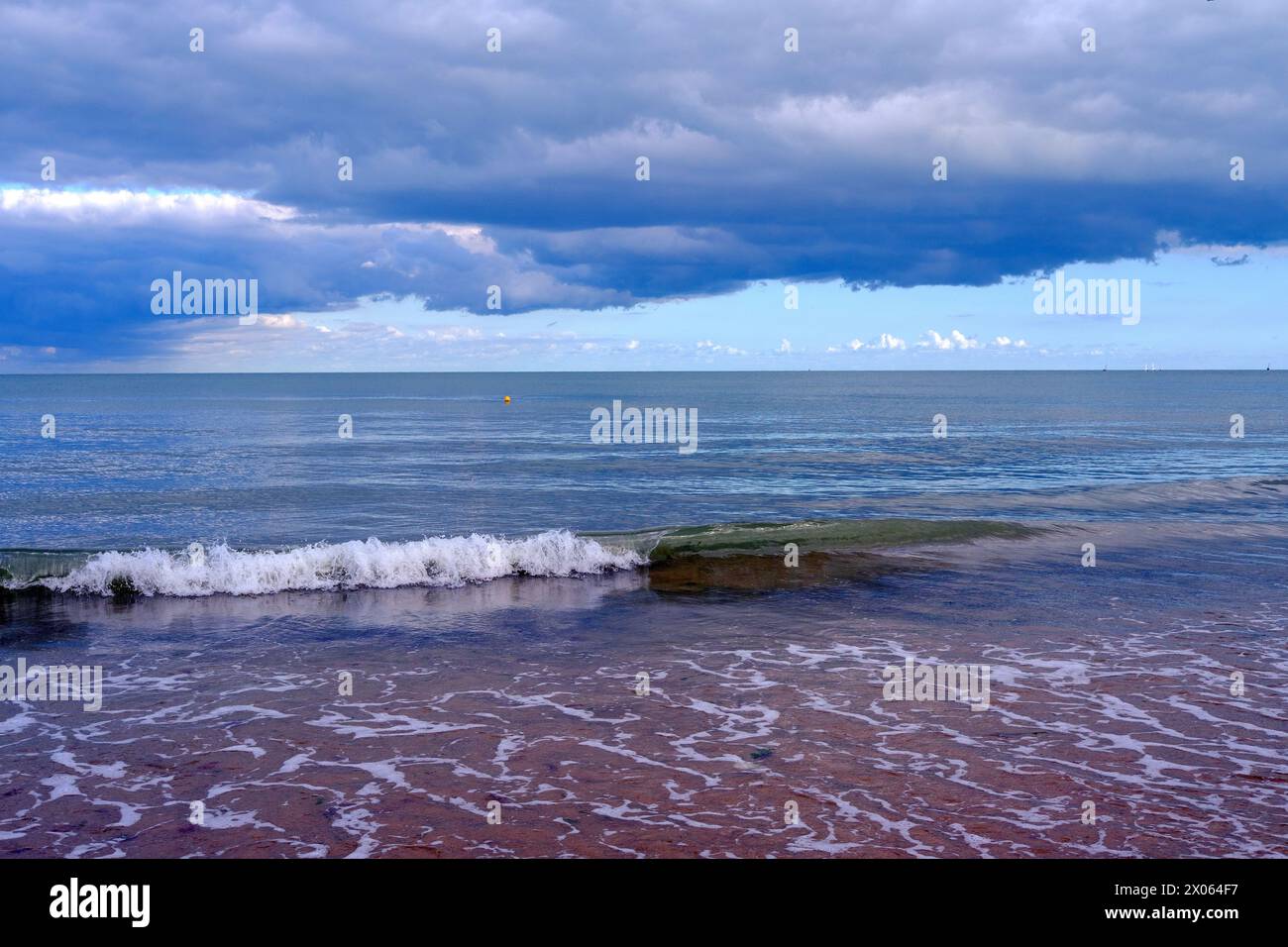 Tranquil beach with calm sea under dark, stormy clouds. Deep blue water, gently lapping the sandy shore. Stock Photo