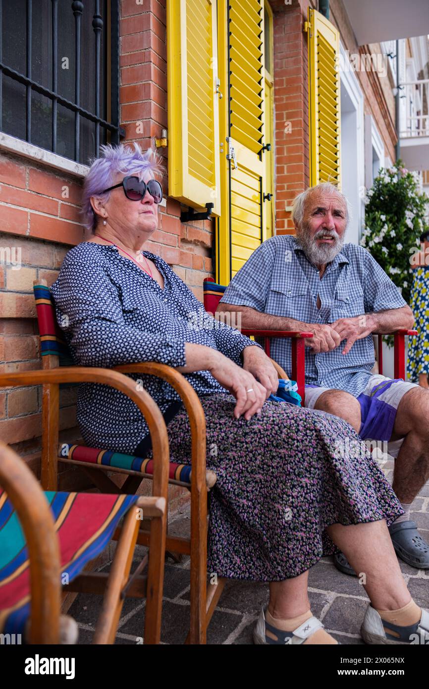 An elderly couple sits on a colorful chair near a brick wall of a house with yellow shutters. Aged woman in sunglasses and man enjoys relaxation while Stock Photo