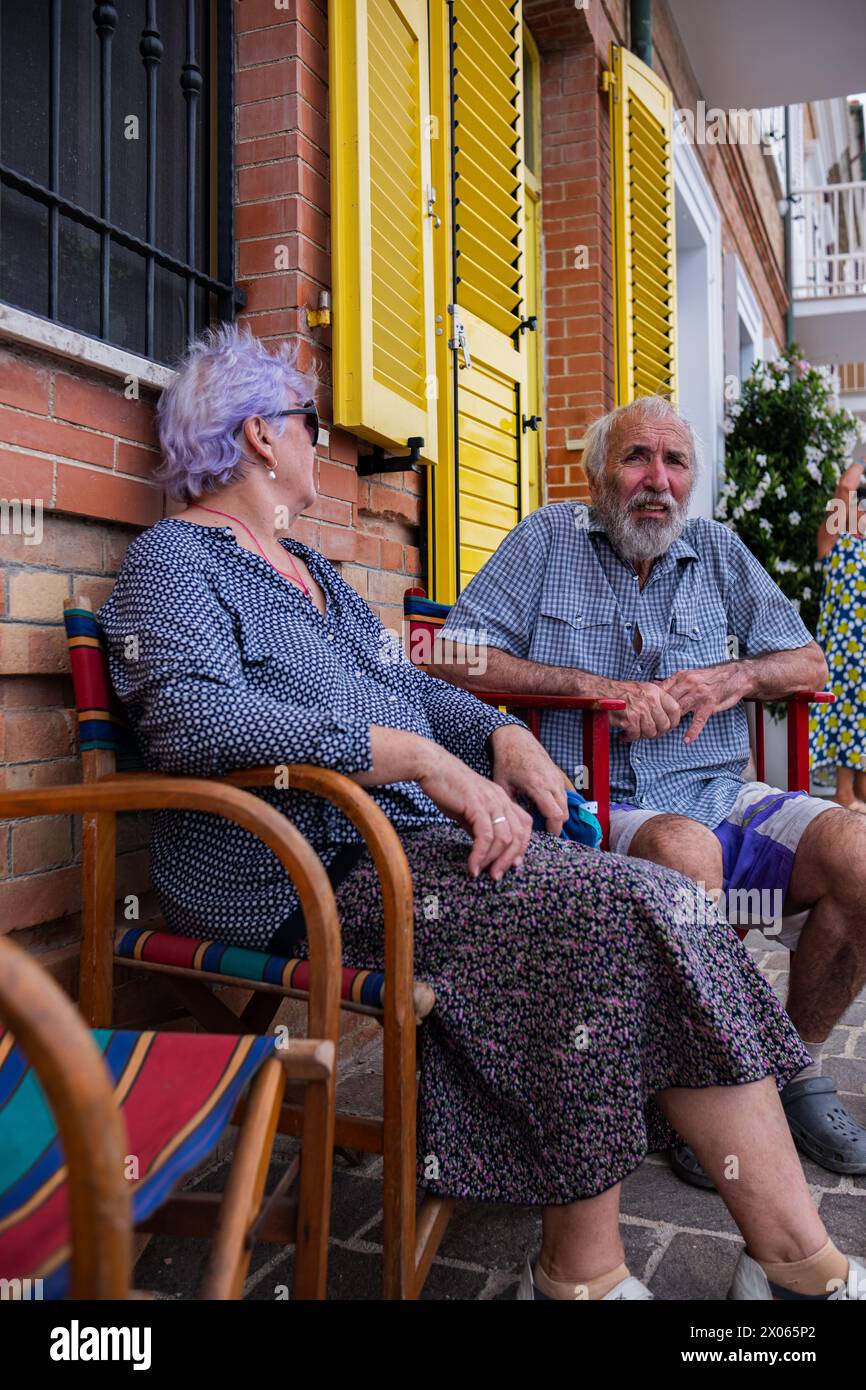An elderly couple sits on a colorful chair near a brick wall of a house with yellow shutters. Aged woman in sunglasses and man enjoys relaxation while Stock Photo