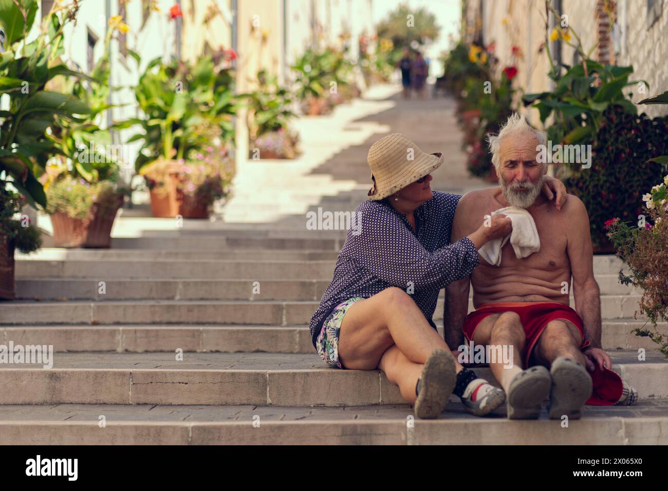 Old travelers are sitting on the steps of a long staircase. A long street with steps is lined with potted plants, an elderly couple smiles while sitti Stock Photo