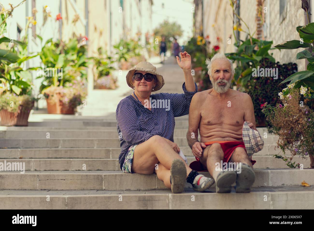 Old travelers are sitting on the steps of a long staircase. A long street with steps is lined with potted plants, an elderly couple smiles while sitti Stock Photo