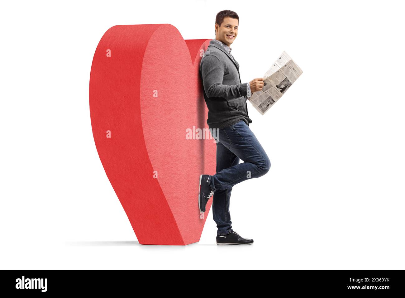 Full length portrait of a guy with a newspaper leaning against a red heart isolated on white background Stock Photo