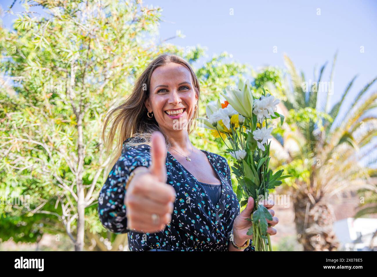 A smiling woman is holding a bouquet of flowers and giving a thumbs up. Stock Photo