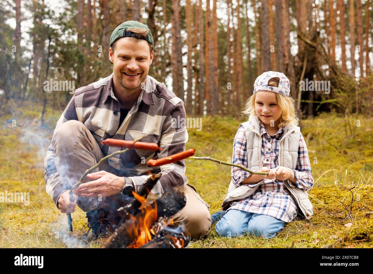 father and daughter spend time together and frying sausages over a bonfire while camping in forest. bonding activities Stock Photo