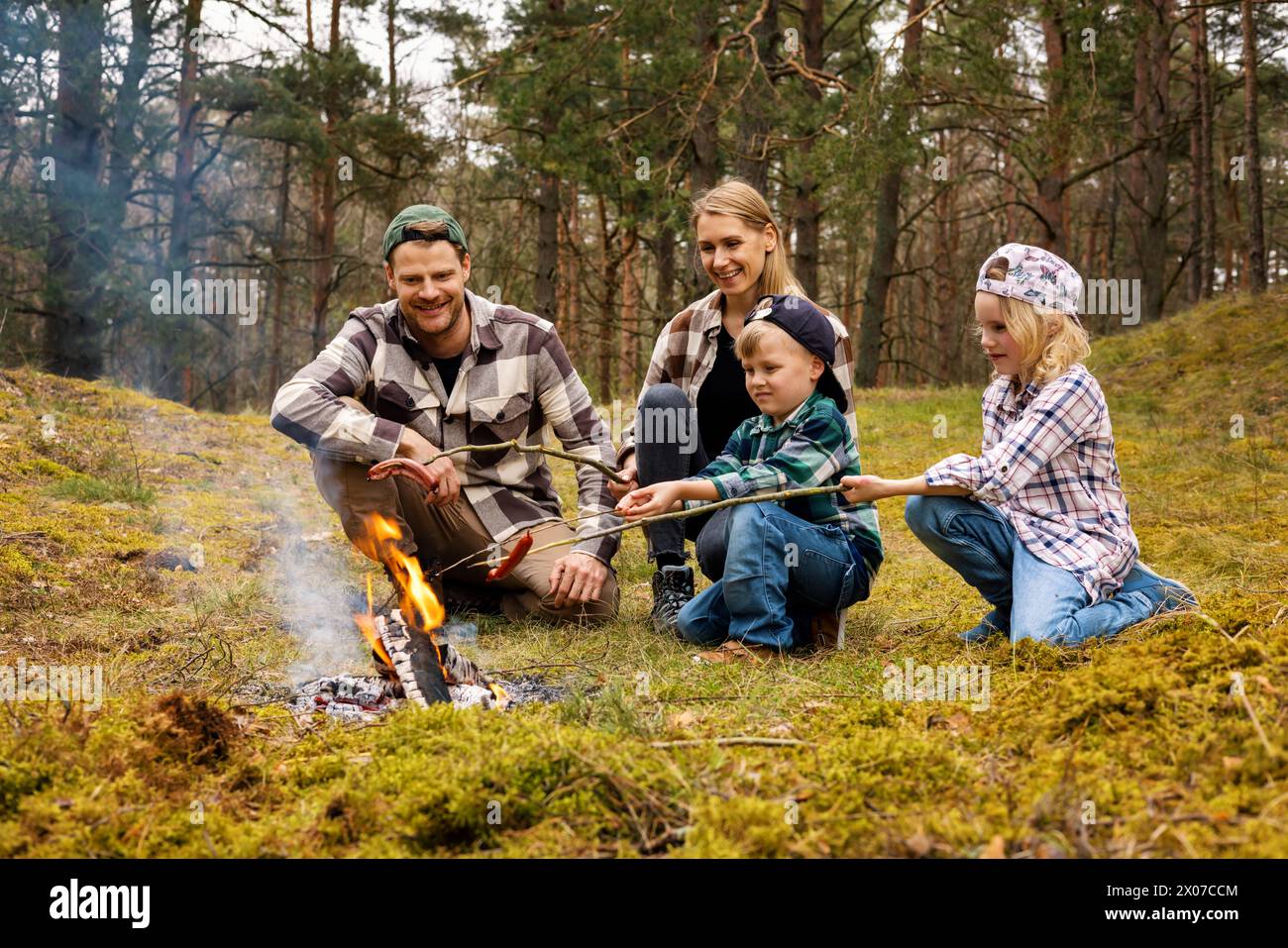 family with two children frying sausages over a bonfire while camping in forest. family time, nature adventure Stock Photo