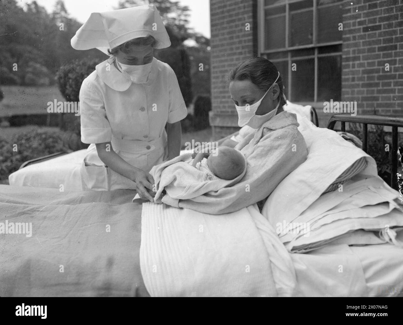 MANSION BECOMES MATERNITY HOME: LIFE AT BROCKET HALL, WELWYN, HERTFORDSHIRE, 1942 - A nurse checks on a mother as she cradles her baby on a bed in the open air outside Brocket Hall. Both women are wearing surgical masks to prevent the spread of germs to the baby Stock Photo