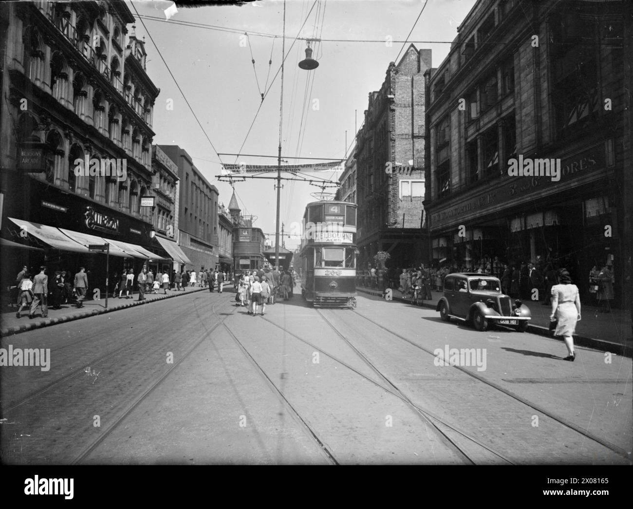 A MIDDLE EAST SOLDIER REVISITS BRITAIN: LIFE IN WARTIME LEEDS, ENGLAND, UK, 1943 - Civilians go about their daily business in a busy scene looking down Briggate in Leeds. On the left hand side, Burton's can be seen alongside the Imperial Hotel. The number 3 tram can also be seen travelling away from the camera, whilst on the right hand side, the number 4 tram is travelling towards the camera on its way to Haddon Place via Roundhay Road. An advertisement for Melbourne Ales is clearly visible on the front of the tram. A banner can just be seen, stretching across the road above the trams, adverti Stock Photo