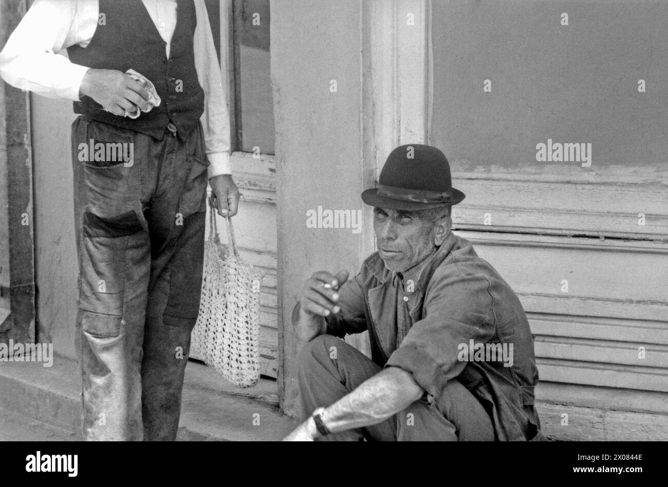 Socialist Republic of Romania, approx. 1974. Man smoking a cigarette while sitting on a sidewalk and chatting with a neighbor out in the street of a small town. Stock Photo