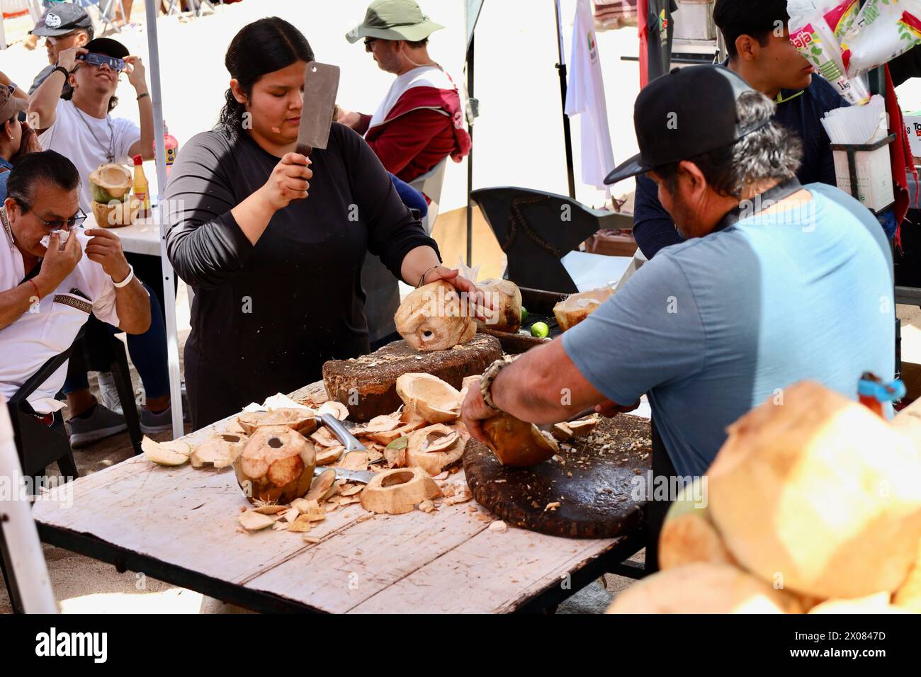 Mazatlan, Mexico. 8th Apr, 2024. A woman with a large cleaver and a man prepare coconuts for refreshments after Eclipse viewing in Mazatlan, Mexico on April 08, 2024. Behind them is a man still using his eclipse glass to view the returning sun. Locals set up make-shift kitchens and tables on the beach, serving this fresh fruit and also fish right from the sea. (Credit Image: © Amy Katz/ZUMA Press Wire) EDITORIAL USAGE ONLY! Not for Commercial USAGE! Stock Photo