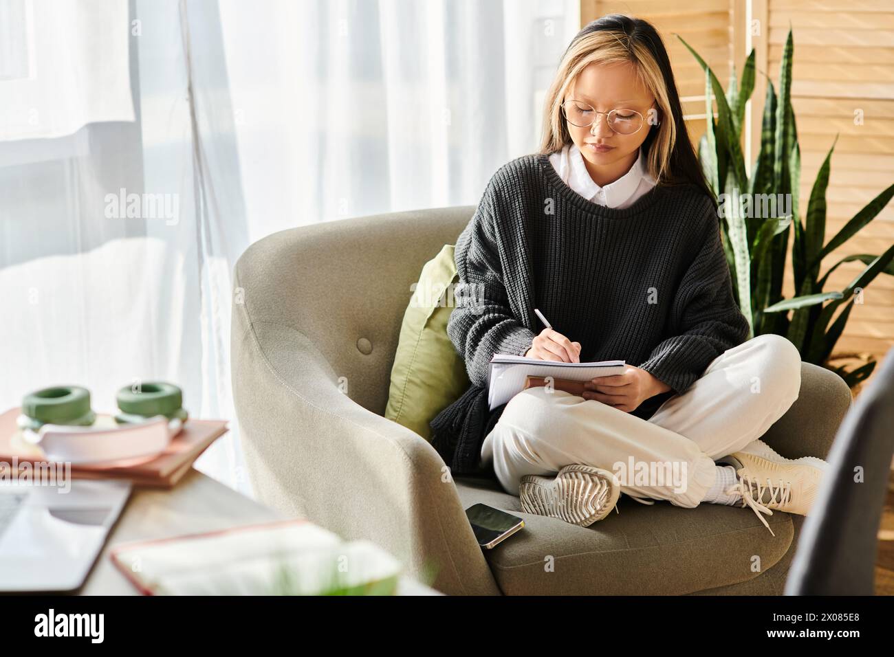 A young Asian woman sits in a chair, engrossed in a book in her lap, embracing the joy of self-education at home. Stock Photo