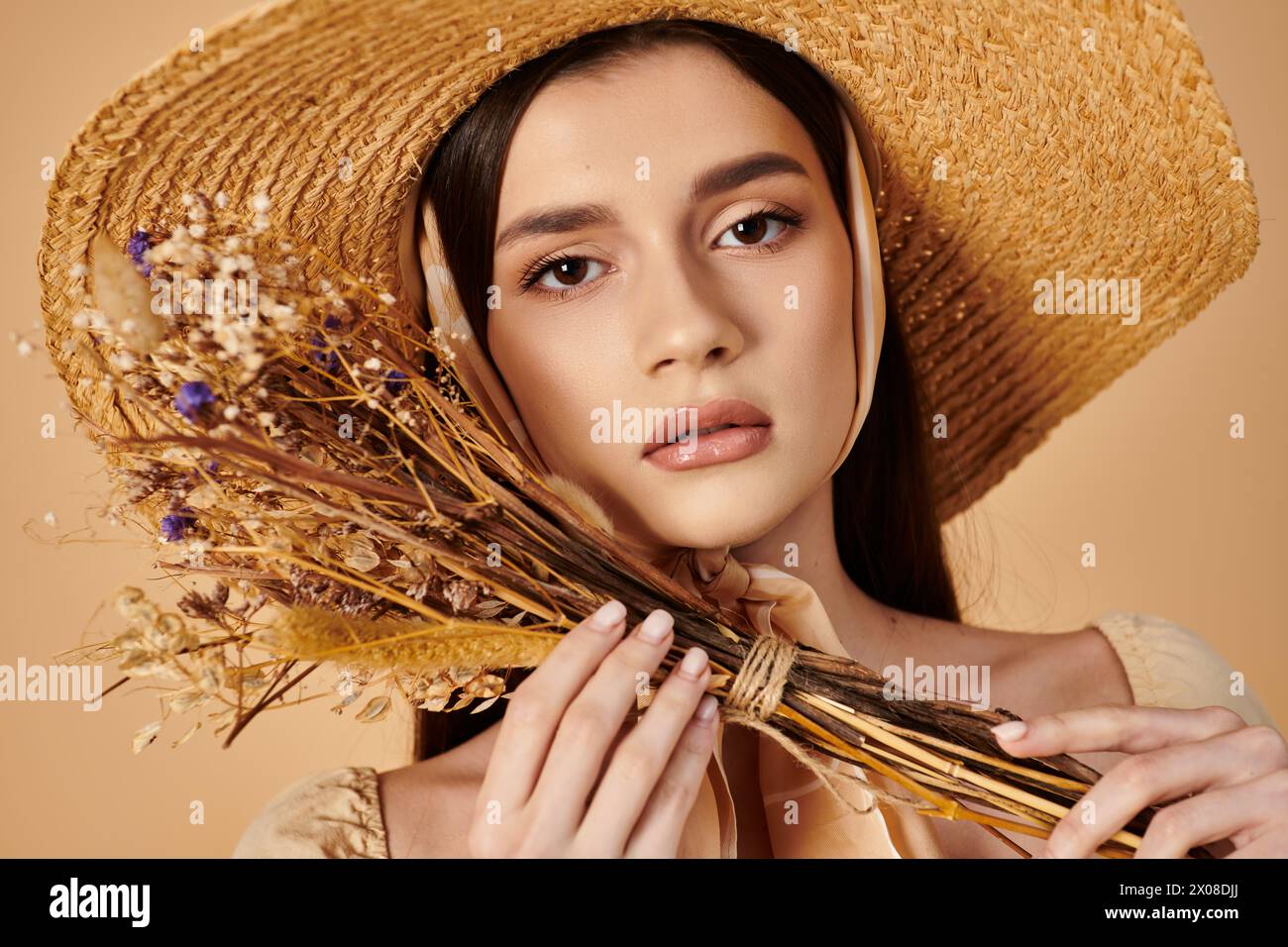 A young woman with long brunette hair smiles softly while holding a bunch of dried flowers in a straw hat. Stock Photo