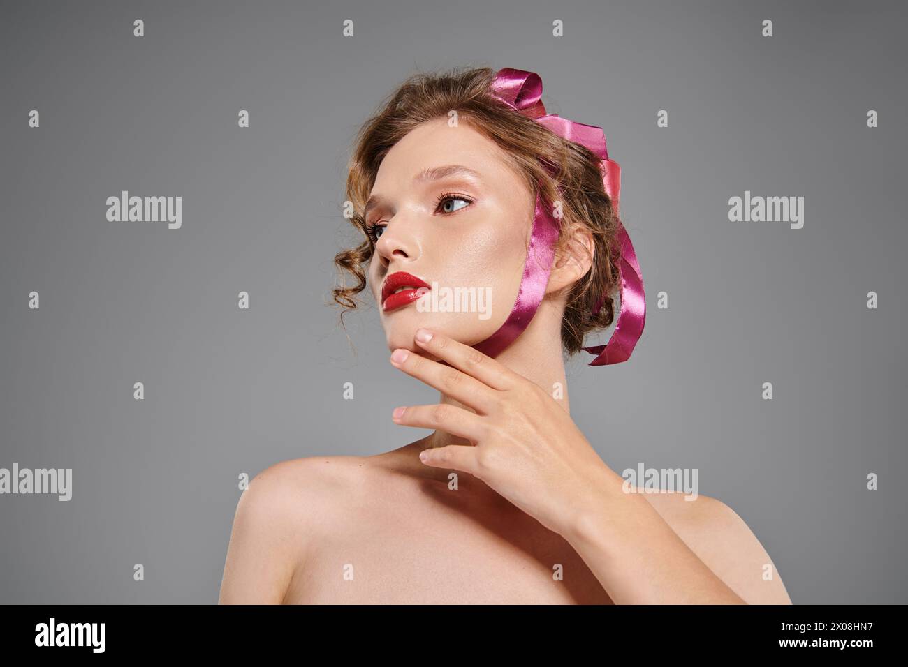 Young woman exudes classic beauty as she confidently poses with a pink bow on her head in a studio setting on a grey background. Stock Photo