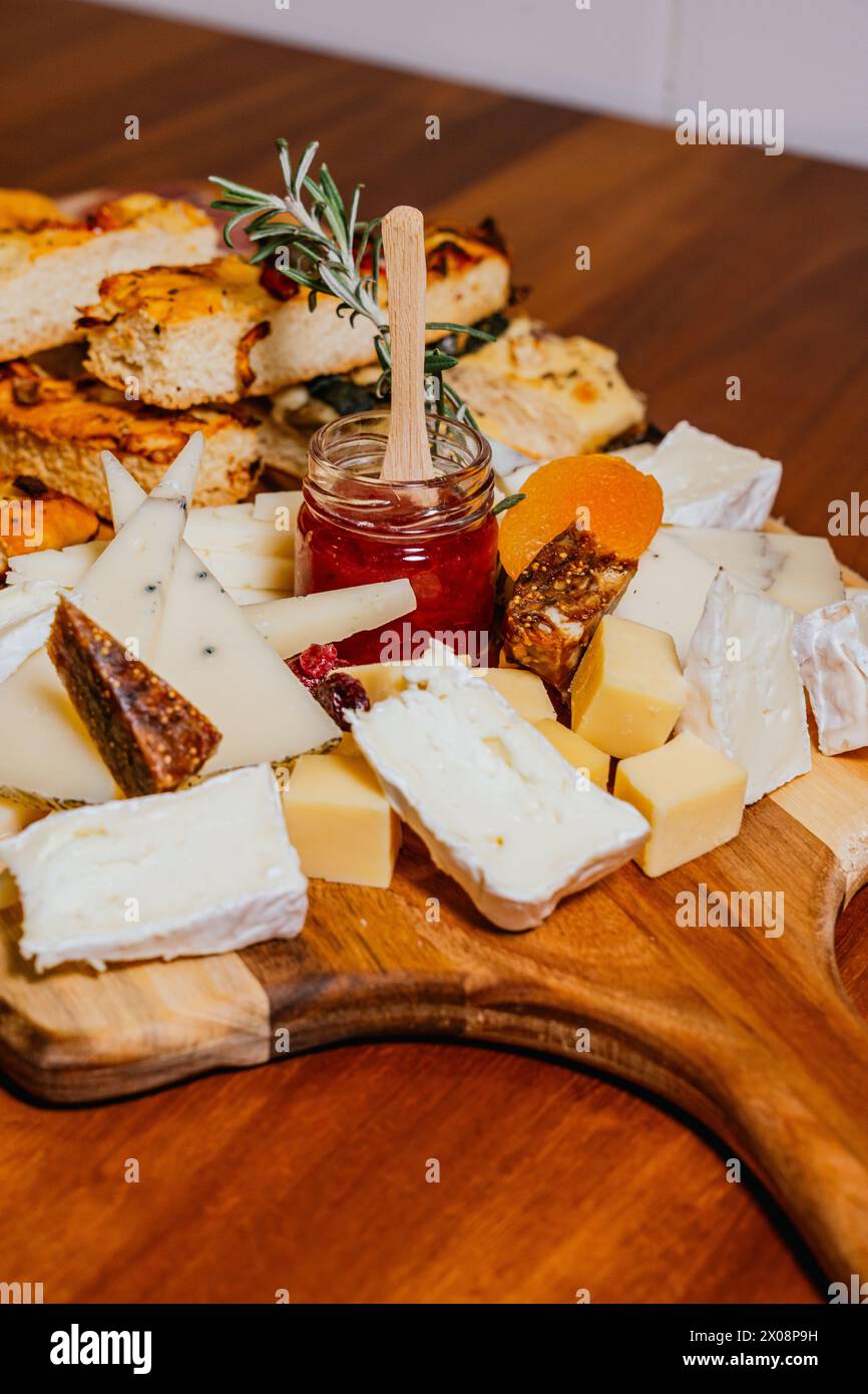 A rustic and inviting cheese platter featuring a selection of fine cheeses, crackers, and a jar of sweet jam, adorned with fresh rosemary Stock Photo