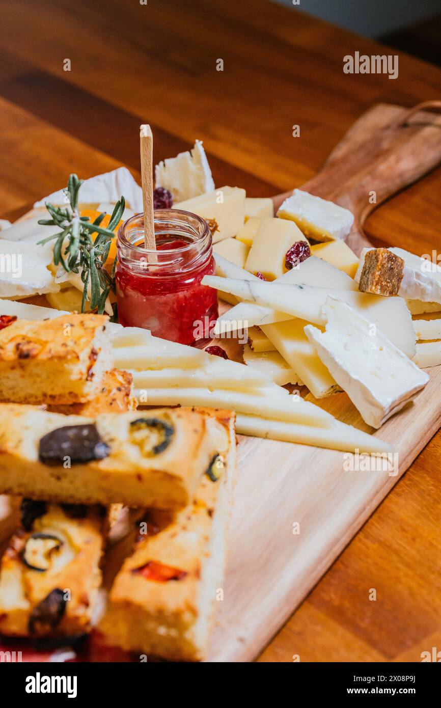 A wooden board featuring an array of artisan cheeses, slices of focaccia, and a jar of berry jam, epitomizing a gourmet snack Stock Photo