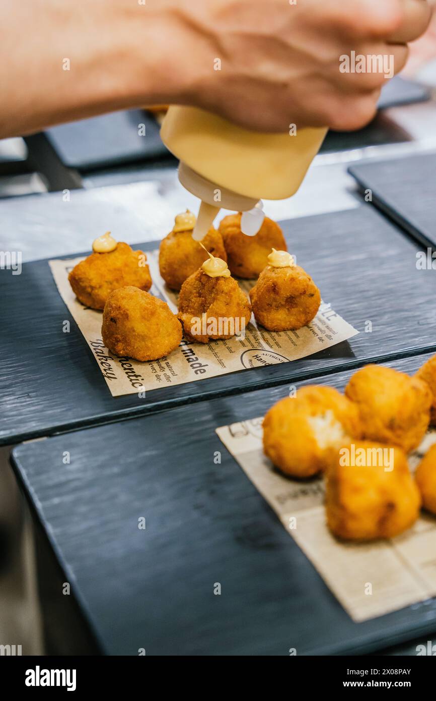 Anonymous chef hands meticulously drizzle a creamy sauce on golden croquettes arranged on a newspaper-lined serving tray Stock Photo