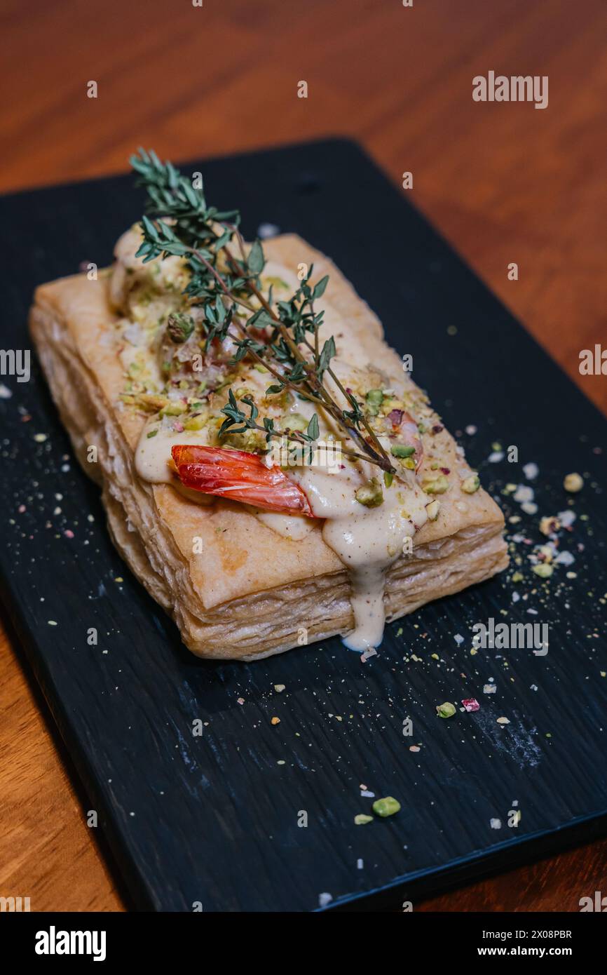 A sumptuous puff pastry dish, garnished with herbs, a succulent shrimp, and drizzled with a creamy sauce, presented on a sleek black slate Stock Photo