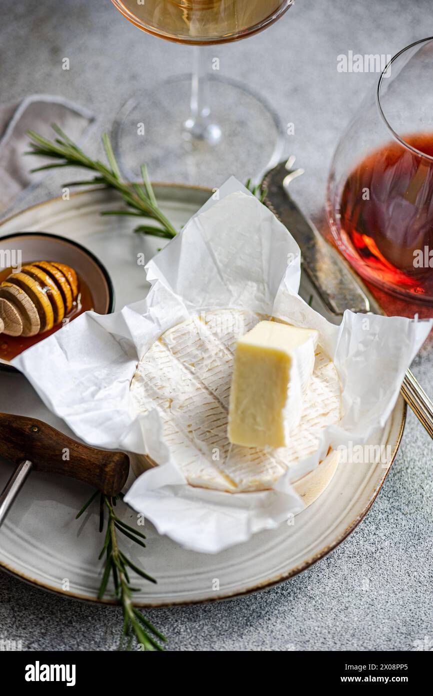 An elegant presentation of brie cheese with honeycomb, rosemary, and wine glasses on a textured surface for sophisticated dining. Stock Photo