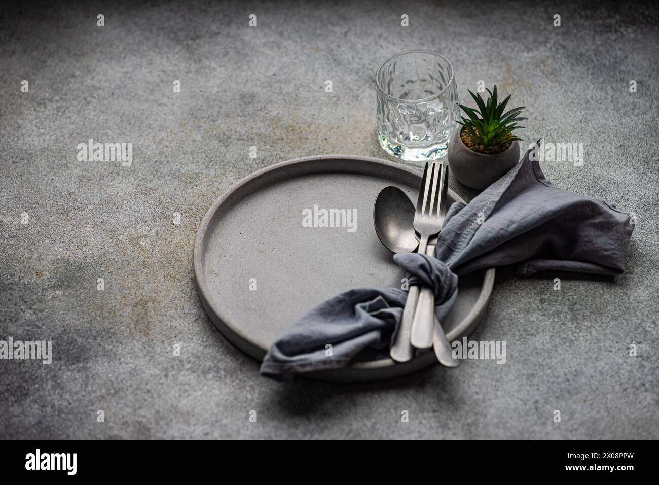 A tasteful minimalist table setting showcasing a ceramic plate, silver cutlery bundled in a grey napkin, a textured glass, and a miniature succulent p Stock Photo