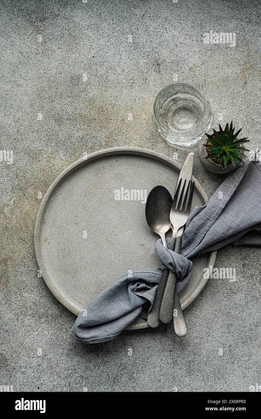 From above of tasteful minimalist table setting showcasing a ceramic plate, silver cutlery bundled in a grey napkin, a textured glass, and a miniature Stock Photo