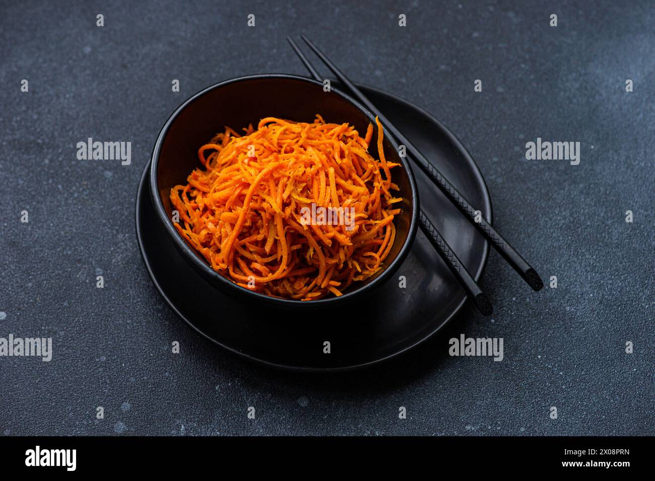 A black bowl filled with shredded carrot salad, accompanied by a pair of black chopsticks, presented on a textured dark gray surface Stock Photo