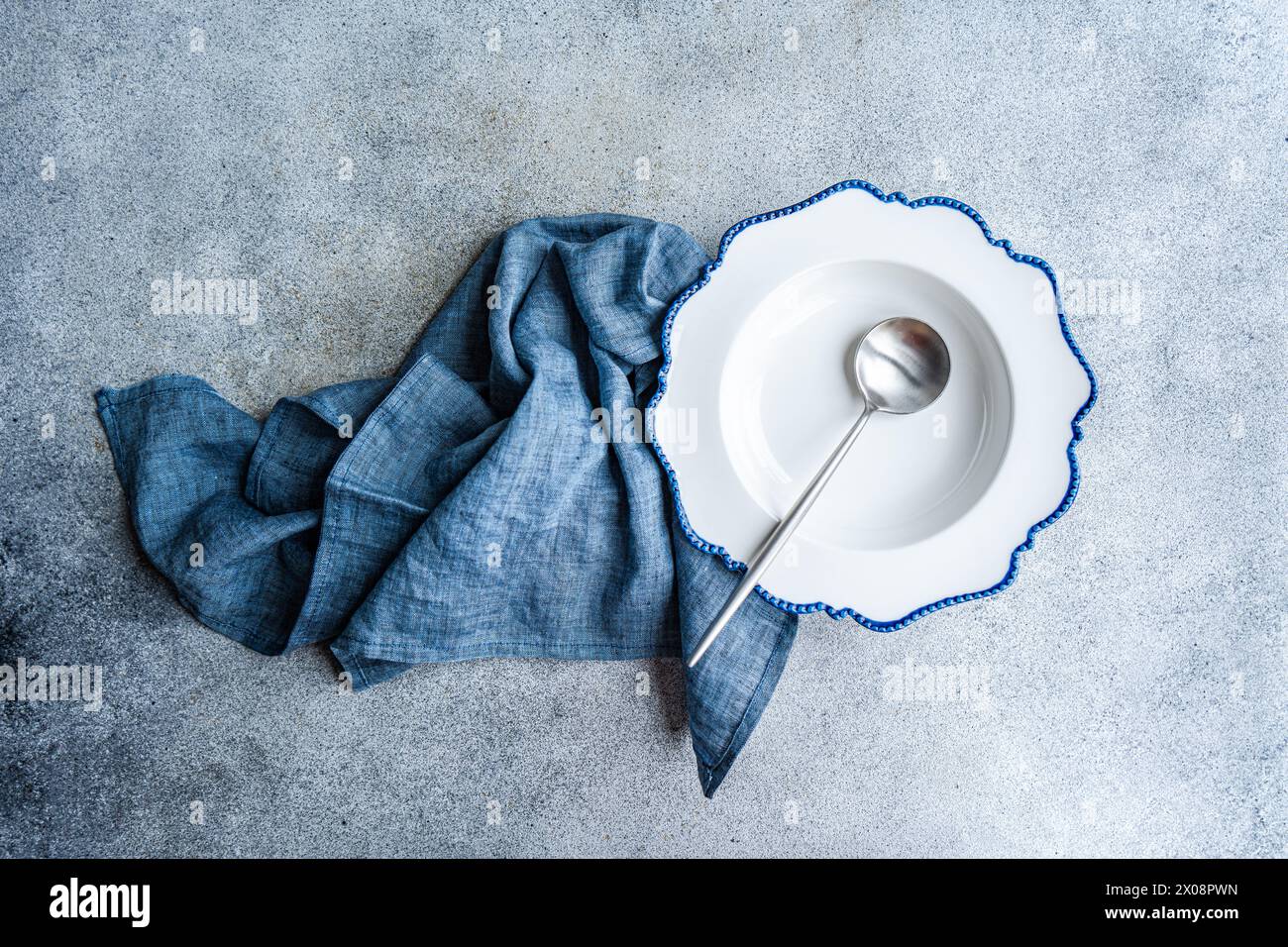 Elegant white plate with blue scalloped edges, a silver spoon, and a blue napkin set on a textured background Stock Photo