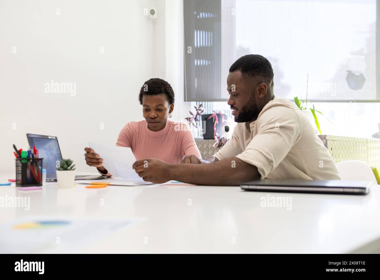 Two African American colleagues analyze documents together at a bright office workspace, with the woman reading a paper and the man focused on a digit Stock Photo