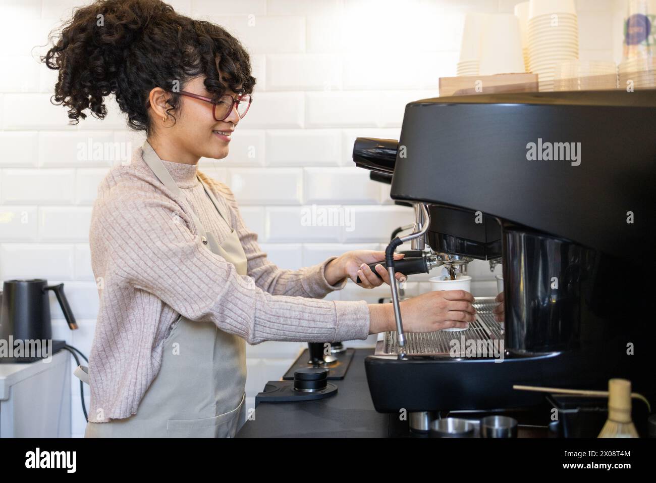 A smiling hispanic female barista expertly operates a professional espresso machine in a bright and modern cafe setting. Stock Photo