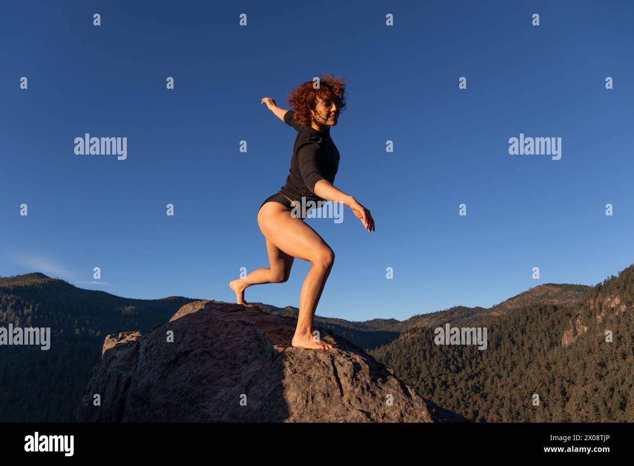 A dynamic image capturing a contemporary dancer striking a poised pose on a rock against a breathtaking mountainous backdrop under a clear blue sky Stock Photo