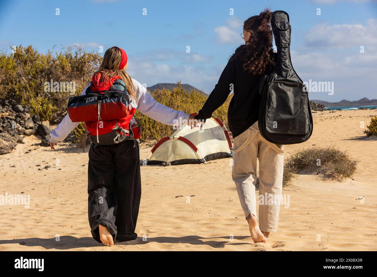 A back view loving couple walks hand in hand near their campsite on the sandy beach, creating memories with a mix of adventure and music Stock Photo