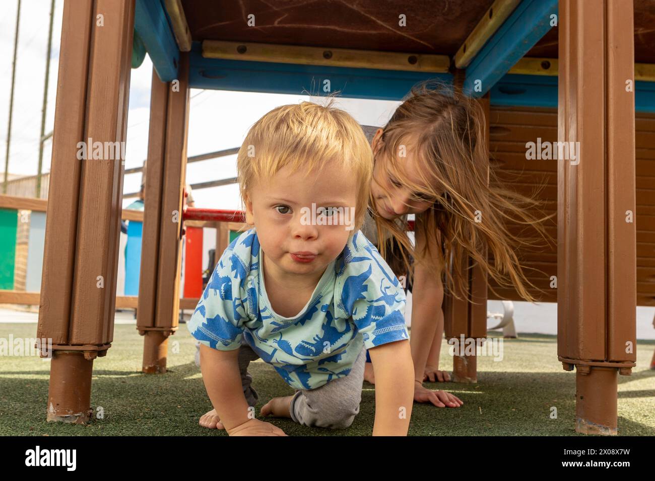 A heartwarming scene with a toddler with Down syndrome and a girl playing under a playground structure, showcasing inclusivity and family bonding in a Stock Photo