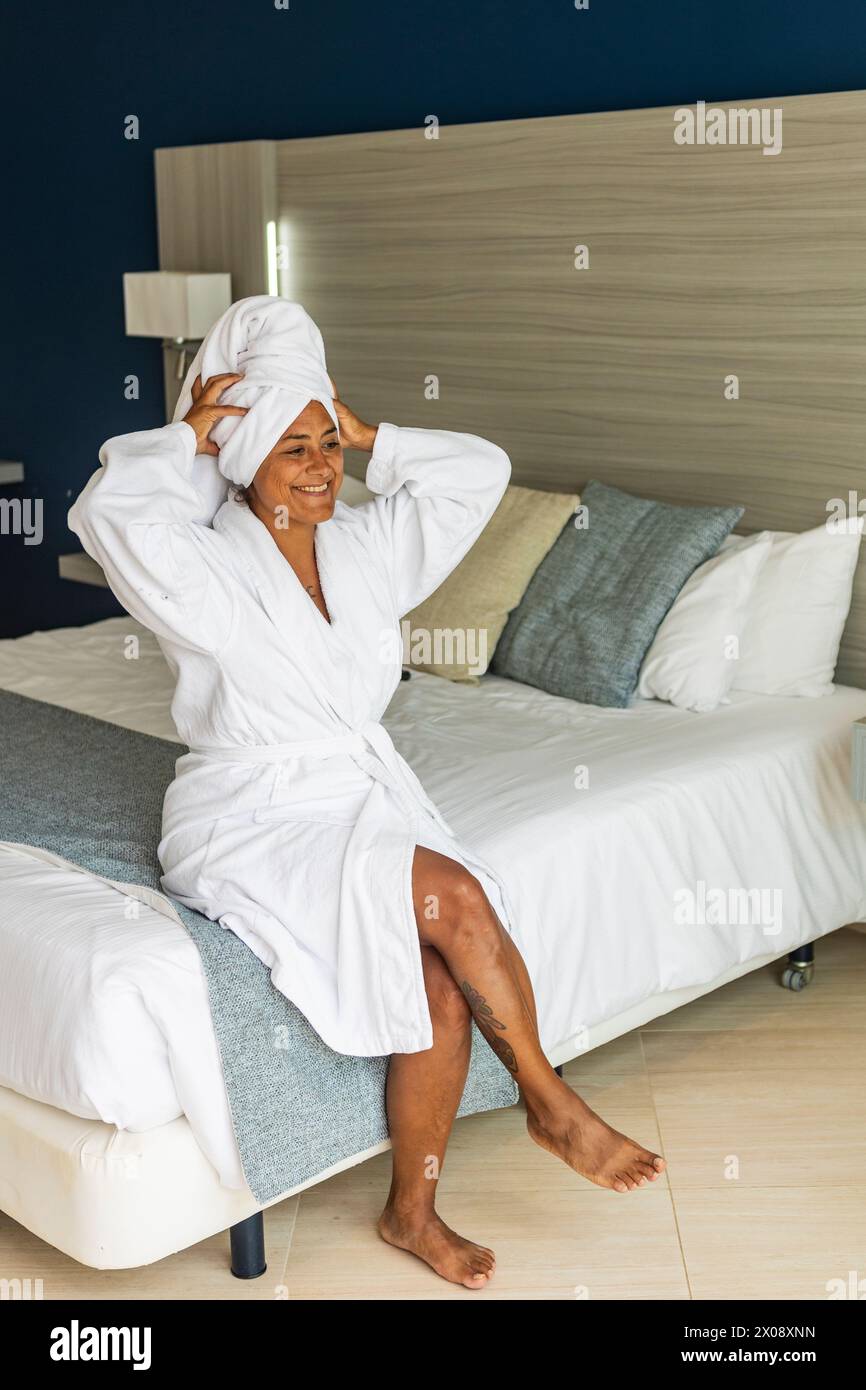 A smiling middle aged hispanic woman in a white bathrobe with a towel wrapped around her head sitting on a hotel bed, radiating relaxation and comfort Stock Photo