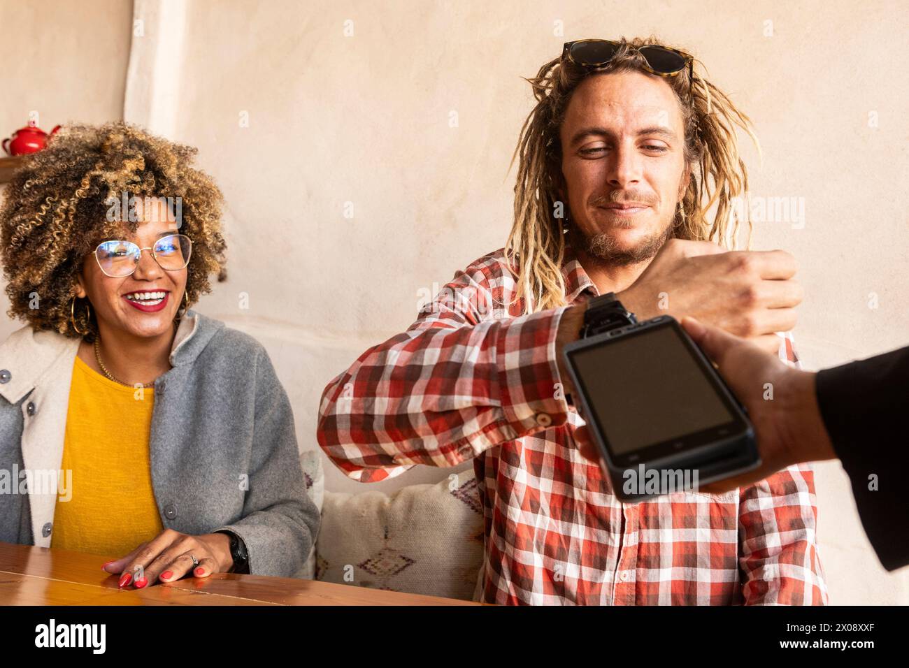 A group of cheerful friends laugh and share a moment of camaraderie as one person uses a digital wristwatch to make a contactless payment at a bar Stock Photo