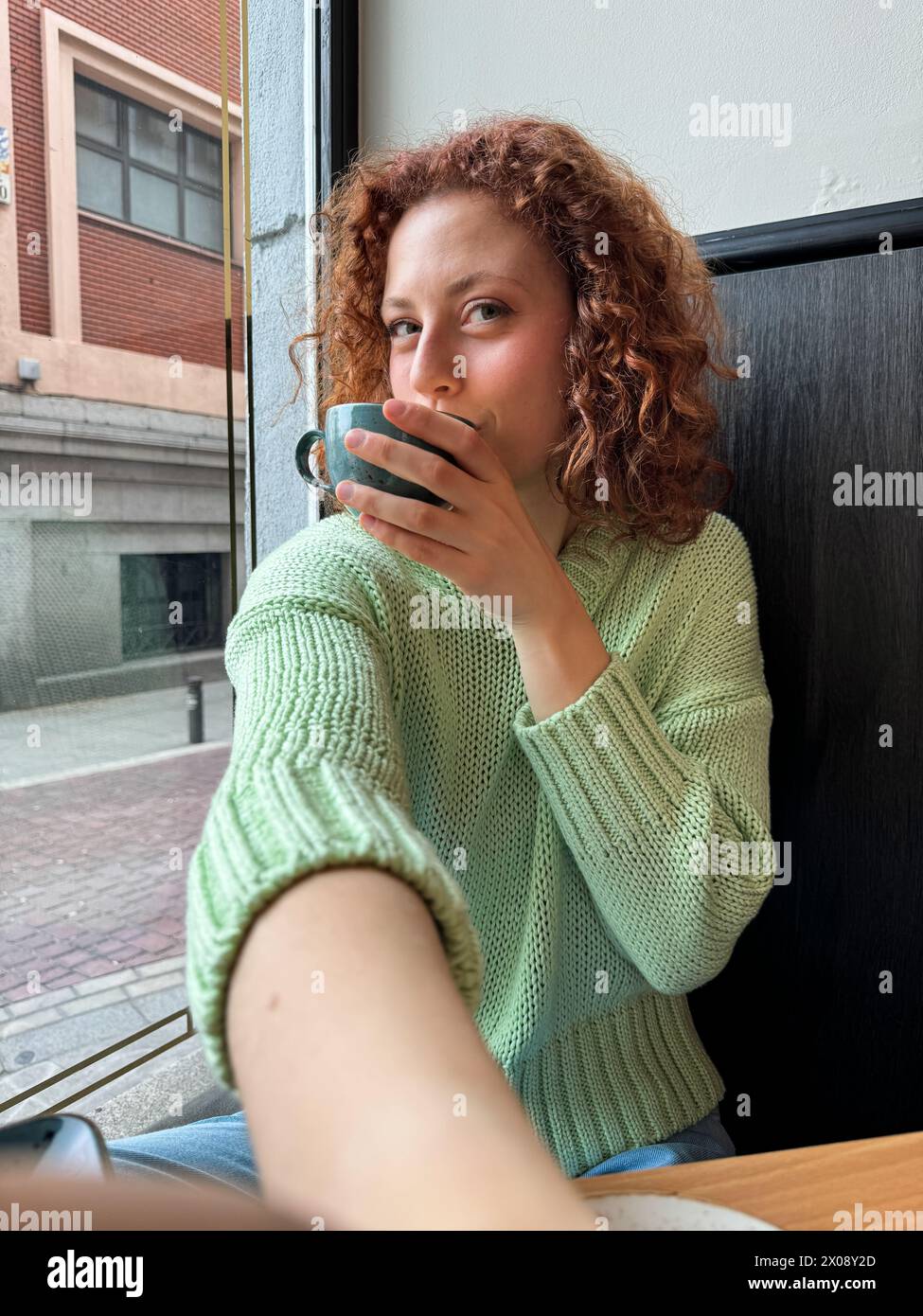 A serene young redhead woman with curly hair enjoys a warm coffee by a window while taking a selfie, offering a sense of calm and reflection Stock Photo
