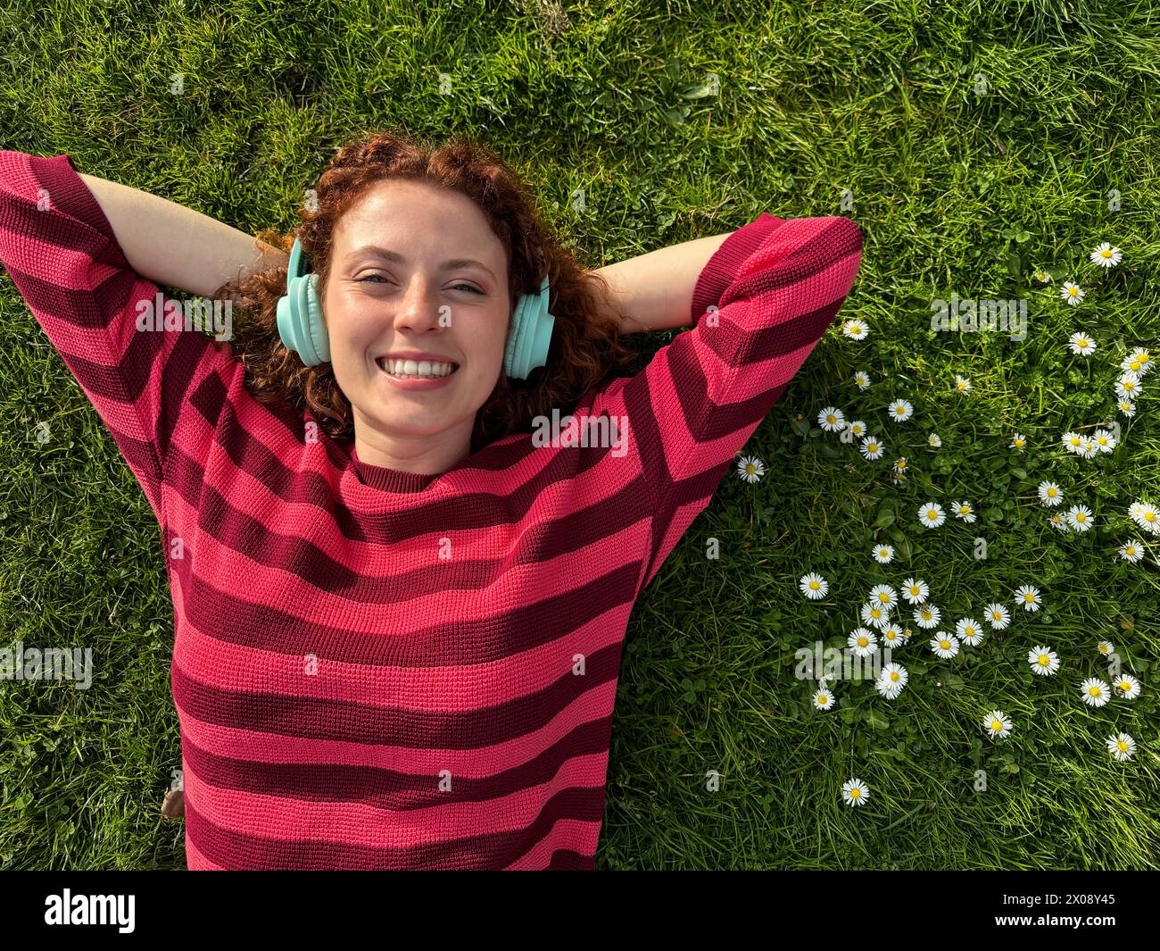 A cheerful young redhead woman lies on the grass with her arms stretched out, enjoying music on
