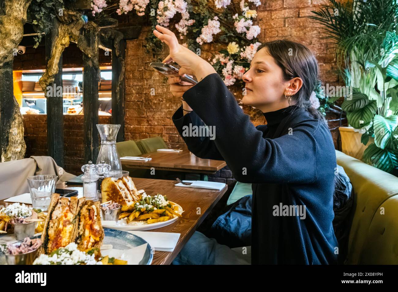 In a quaint restaurant setting, a content diner snaps a picture of a tempting sandwich with her phone, surrounded by lush foliage decor Stock Photo