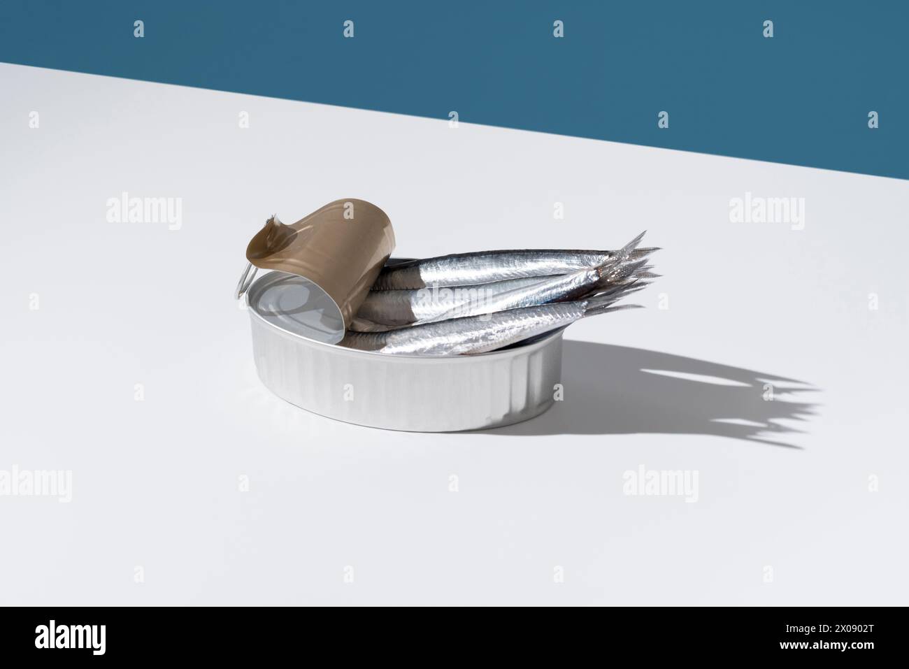 Gleaming fresh anchovies overflow from an unlidded tin can, set against a sharp blue and white divide, casting playful shadows Stock Photo