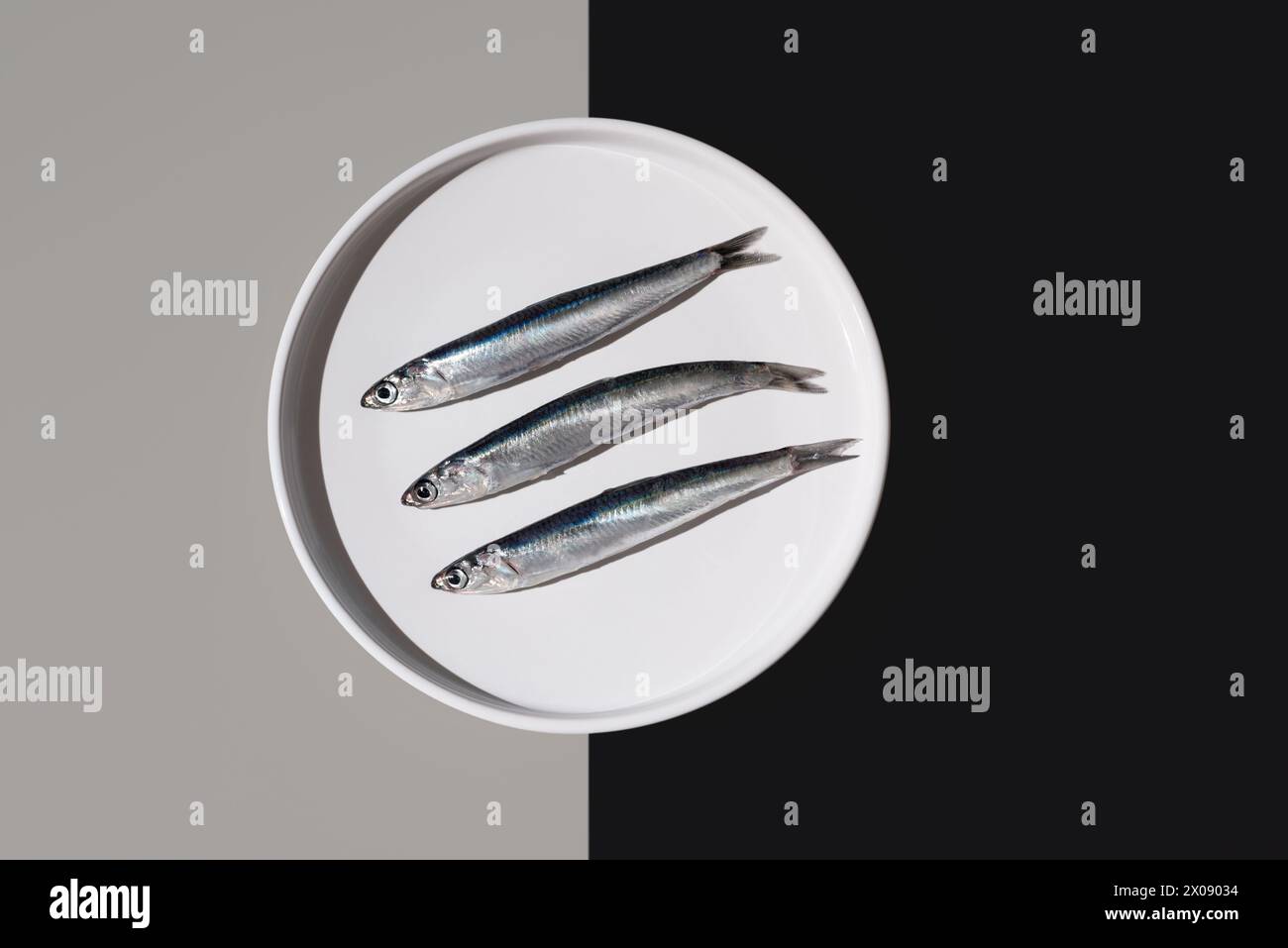 From above of three silvery fresh anchovies aligned on a white round plate, artfully divided by a yin yang background of black and gray Stock Photo