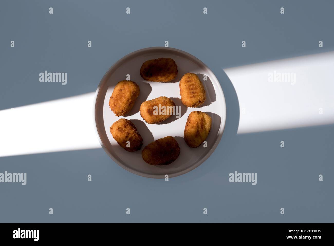 A ceramic plate filled with Spanish appetizer croquettes is partially illuminated by sunlight, creating a dynamic contrast between light and shadow on Stock Photo