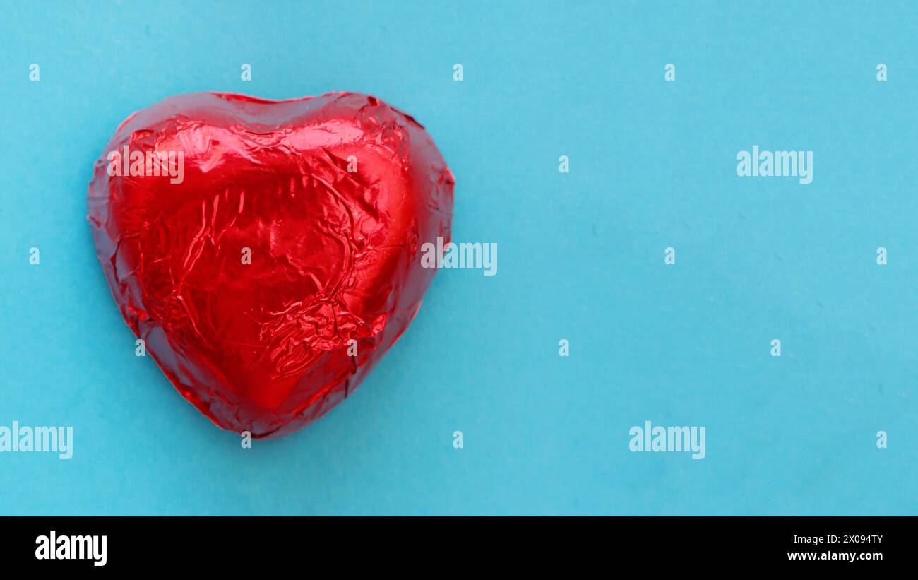 Large heart-shaped candy in red foil on a blue paper background. Flat lay. Stock Photo