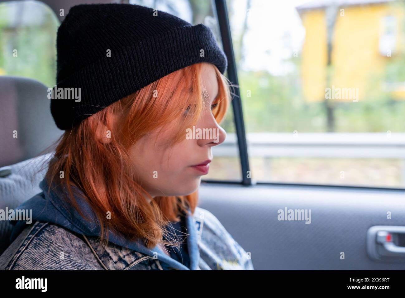 A pretty girl with pink hair in a black knitted hat in the back seat of a car, looking out the window, she is immersed in thoughts about her future, a Stock Photo