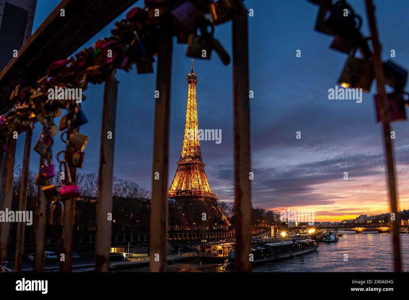 The Eiffel Tower seen between the metal bars of a fence filled with love locks shines against the night sky, casting a warm glow on the Parisian city. Stock Photo